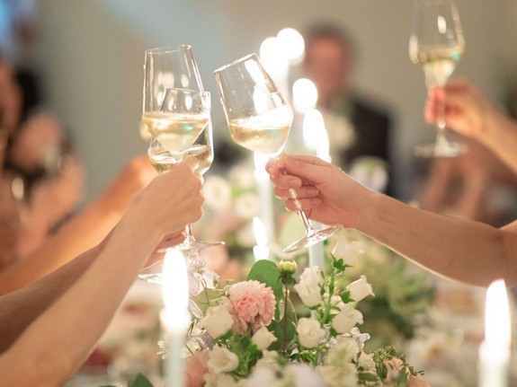 Mehrere Personen stoßen über einer mit Blumen und Kerzen geschmückten Tafel mit Weißwein an.