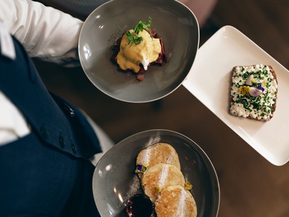 Ein leckeres Frühstück für den perfekten Start in den Tag Ein Servierer bringt ein köstliches Frühstück mit Pfannkuchen, einem herzhaften Brotbelag und einer Portion Eier Benedict zum Tisch.